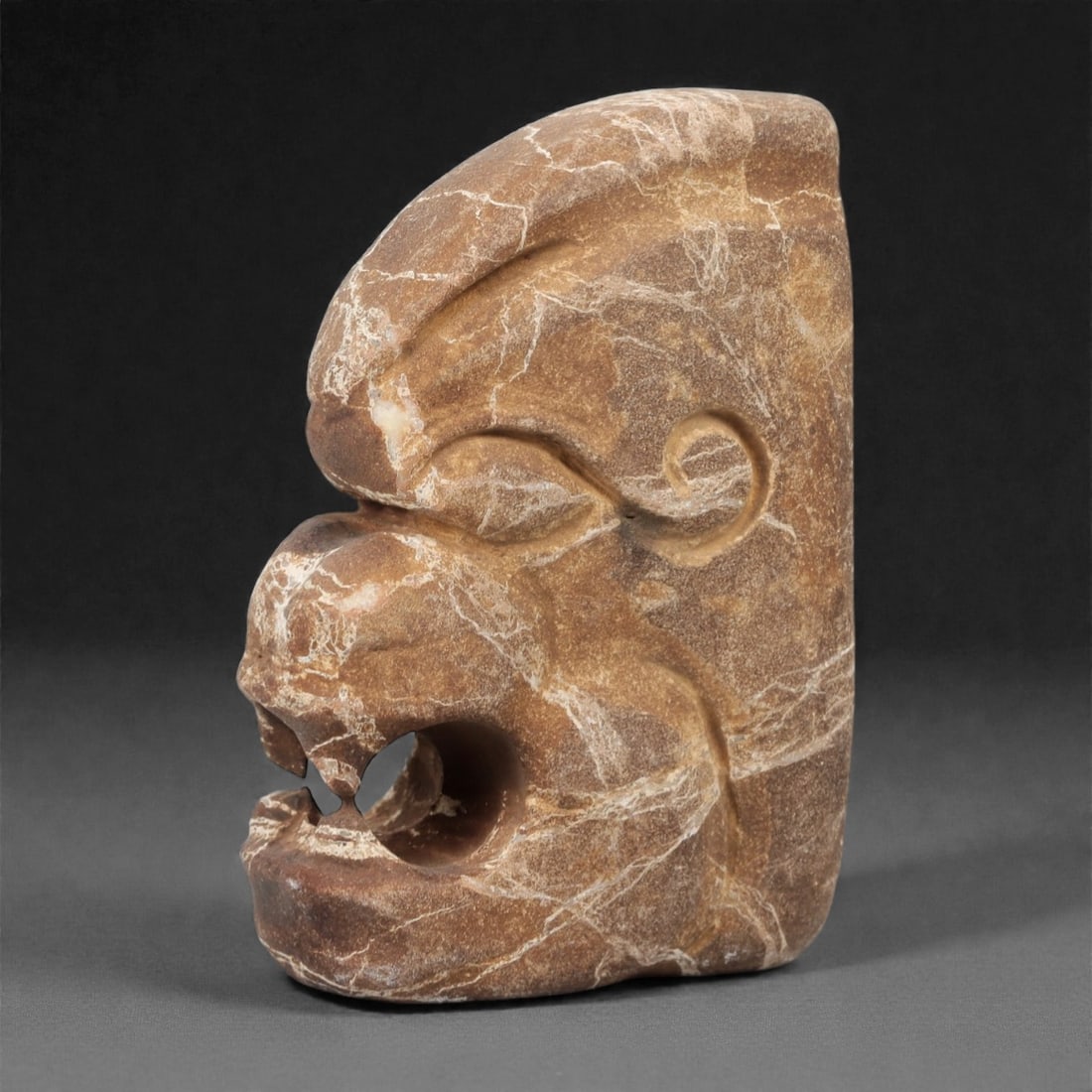 A Pre-Columbian Stone Zoomorphic Head, 10th-15th Century, 31FM: Carved from brown stone with prominent white veining, the object is shaped as a zoomorphic head with a fierce expression, open mouth, and stylized facial features. Incised lines define the eye socket,
