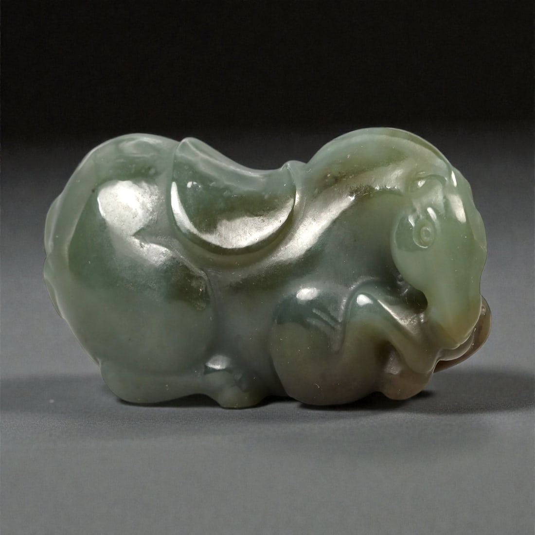 A Chinese Jade Horse Figure, 20th Century, 86FA: Carved from translucent green jade with areas of darker mottling and subtle tonal variation, the figure is worked in the round as a recumbent horse shown resting with its legs tucked beneath the body.