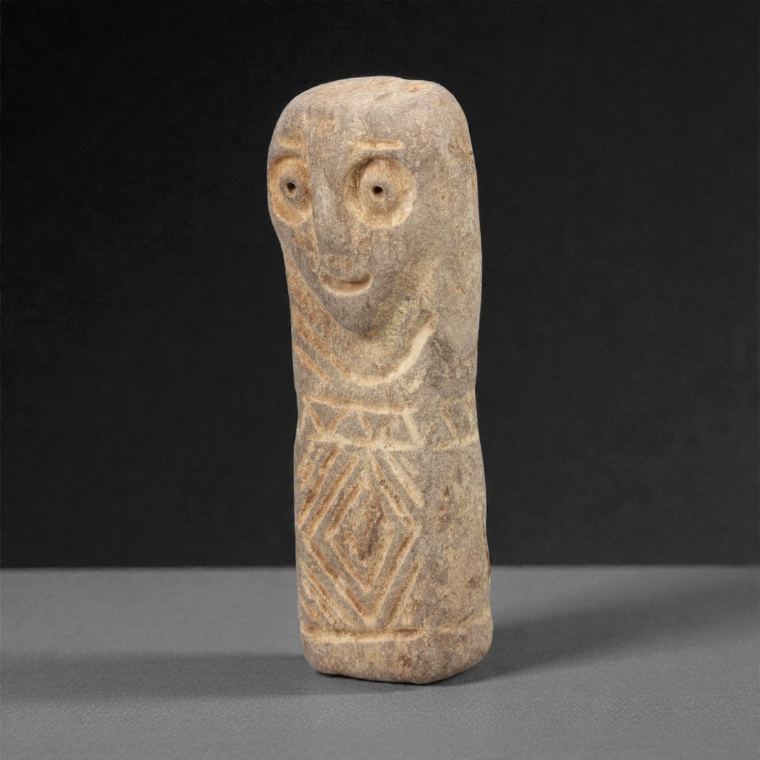 A Near Eastern Stone Idol, 3rd-2nd Millennium BCE, 79FL: Carved from light brown stone with natural surface patination, the object is formed as a slender, standing humanoid idol. The head is rendered in low relief with prominent, perforated circular eyes, a