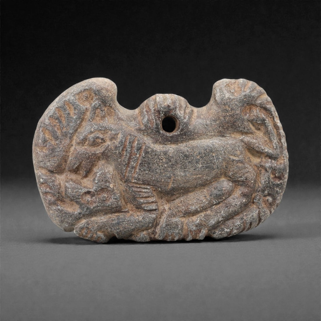 A Near Eastern Schist Amulet, 3rd-2nd Millennium BCE, 90FL: Carved from grey schist with natural surface patination and reddish-brown incised decoration, the object is formed as a flattened, irregularly shaped amulet with a central perforation for suspension.