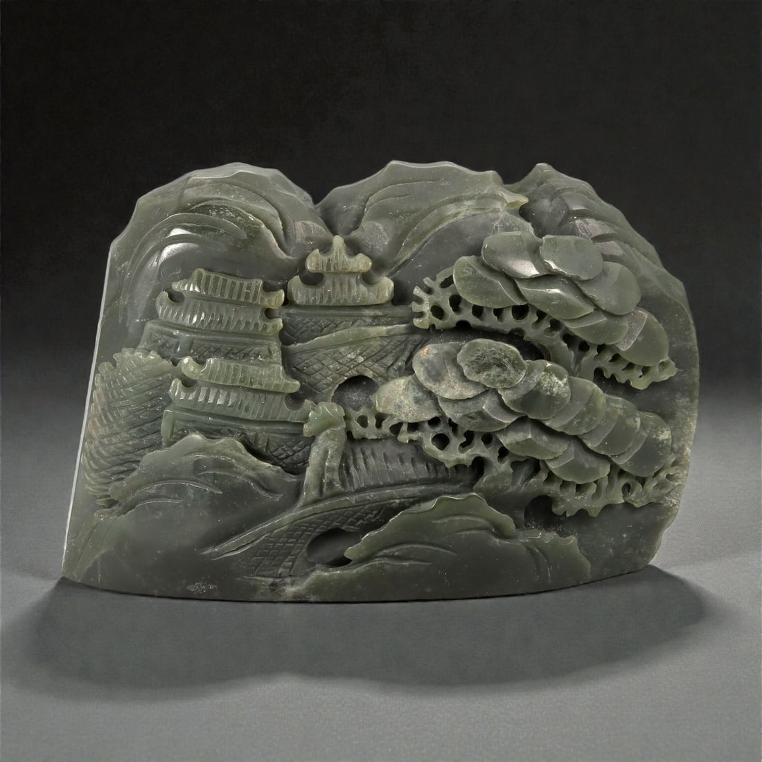 A Chinese Nephrite Jade Landscape Display, 20th Century, 15FD: Carved from dark green Hetian nephrite jade with subtle tonal variation and scattered natural inclusions, the sculpture is worked as a freestanding landscape composition in high relief. The scene depi