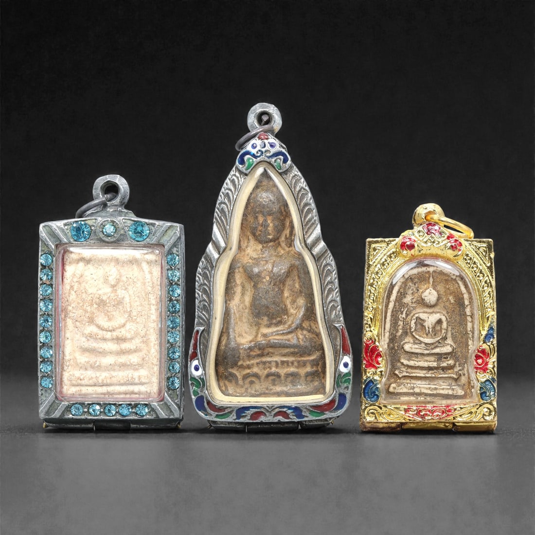 A Group of Three Thai Terracotta Buddha Amulets, 20th Century, 88XJ: Each amulet is formed from terracotta, enclosed in a metal case with enamel and glass accents, and fitted with a suspension loop for wear. The left and right amulets depict a seated Buddha in bhumispa