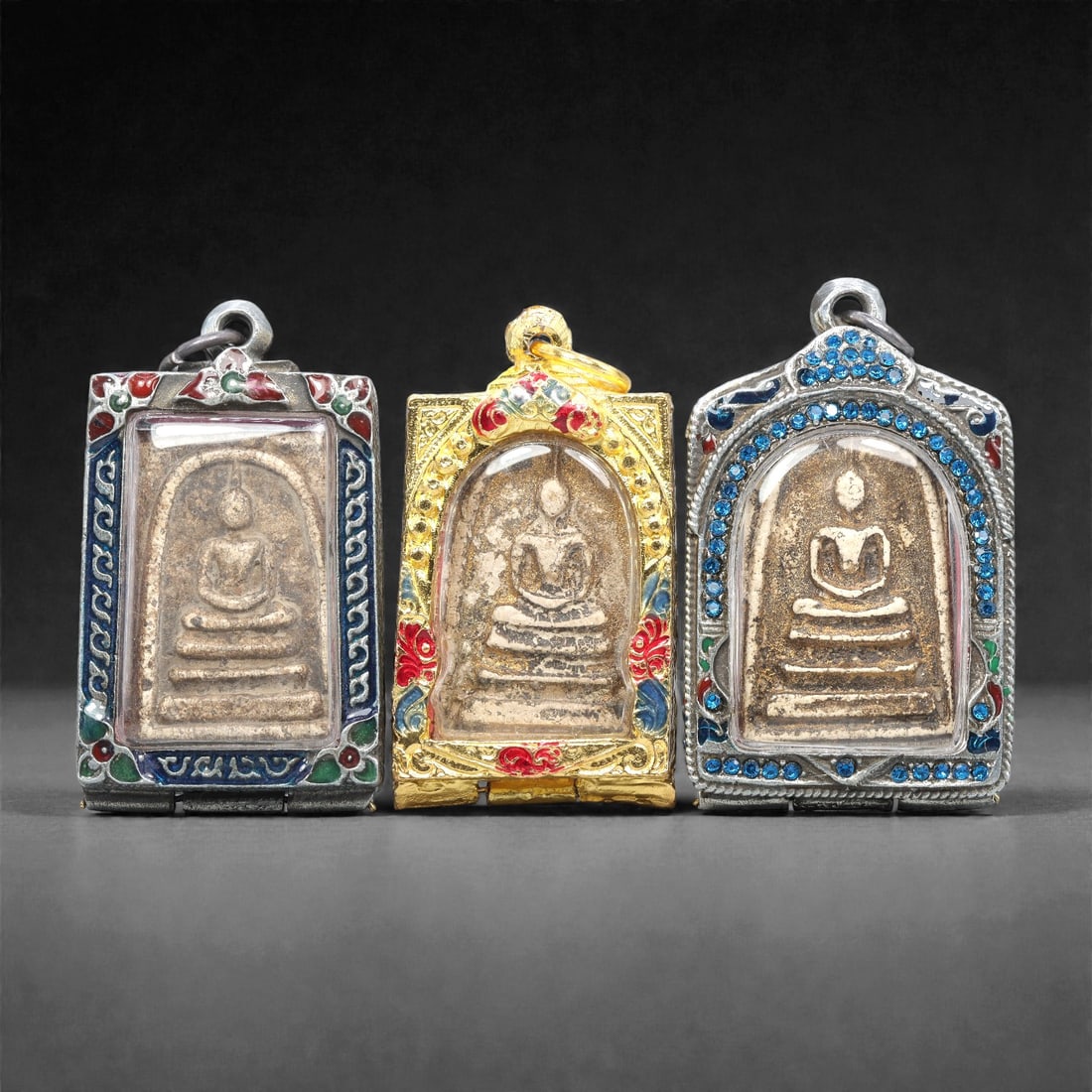 A Group of Three Thai Terracotta Buddha Amulets, 20th Century, 86XJ: Each amulet is formed from terracotta, enclosed in a metal case with enamel and glass accents, and fitted with a suspension loop for wear. The amulets depict a seated Buddha in bhumisparsha mudra, wit