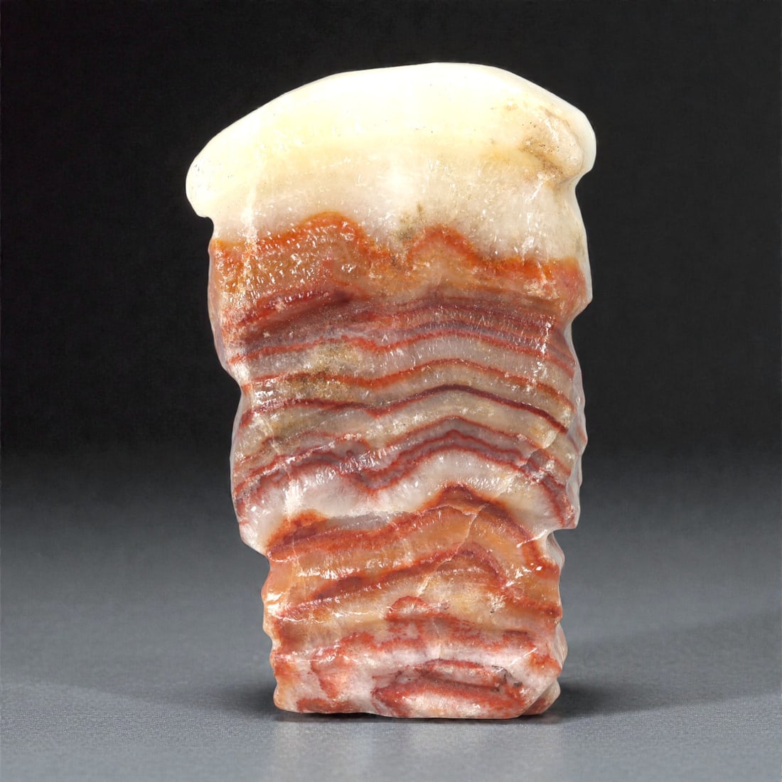 A Chinese Stone Carving Imitating Pork, 20th Century, 35FC: Carved from banded stone with pronounced stratified layers of cream, pale grey, and reddish-brown tones, the object is fashioned to resemble a slice of cooked pork. The form is vertically oriented, wi