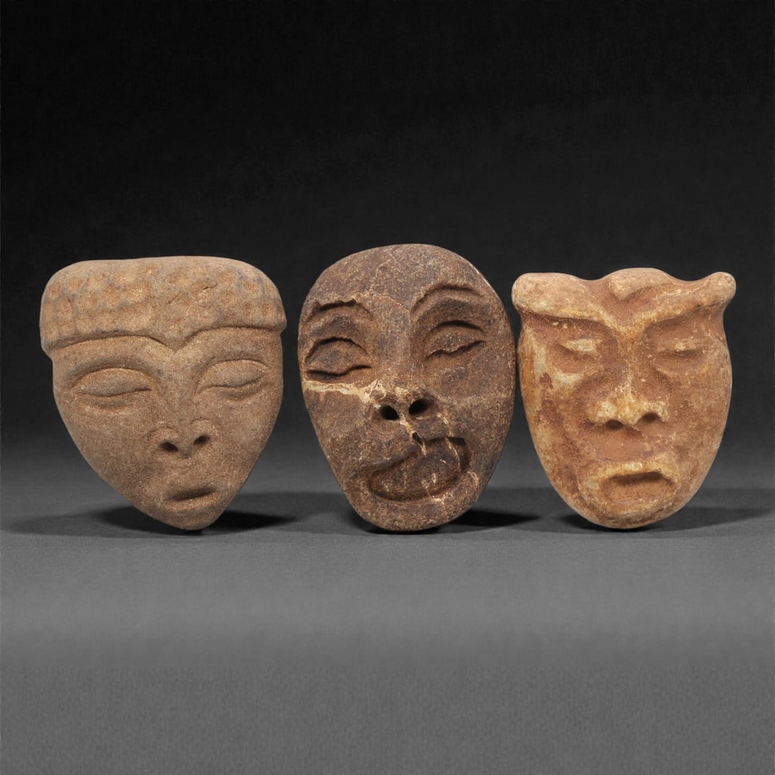 A Group of Three Medieval Stone Maskette Amulets, 10th-14th Century CE, 7ZH: Comprising three distinct maskette amulets, each carved from stone with natural color and texture variations. The first, of light brown stone with a granular surface, is teardrop-shaped with closed ey