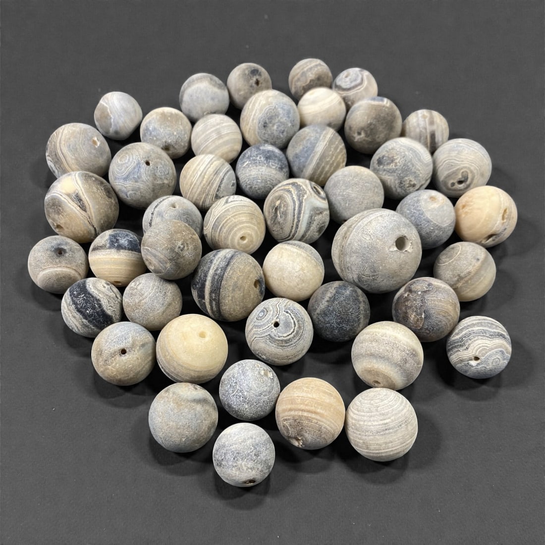 A Group of Tibetan Agate Beads, 18th-19th Century, 62V: Comprising forty-six spherical and near-spherical beads carved from agate, the group presents a cohesive range of cool grey, blue-grey, cream, and pale beige tones, with many examples exhibiting finel