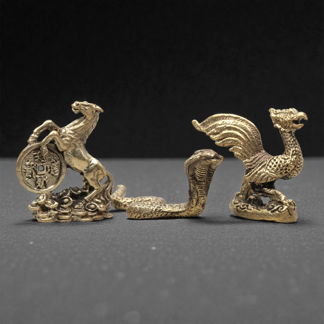 A Set of Three Thai Brass Miniature Figures, 20th Century, 72ZG: Cast from polished brass with a warm golden patina, the set comprises three distinct miniature figures: a rearing horse with a coin, a coiled cobra, and a standing phoenix. The horse is depicted in a