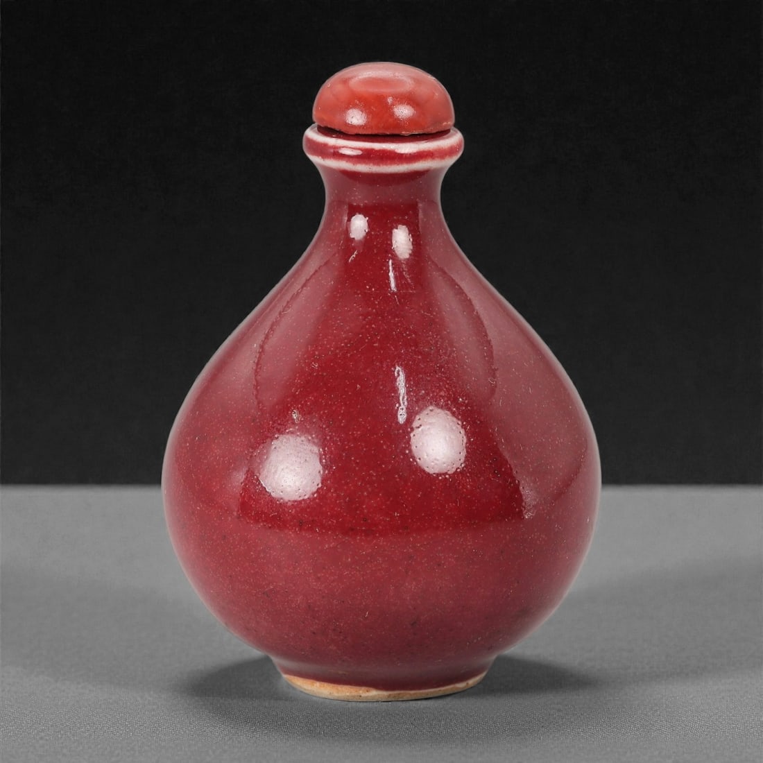 A Chinese Glazed Ceramic Snuff Bottle, 20th Century, 92FG: Thrown from a ceramic body and covered with a deep red glaze with fine speckling and subtle tonal variation, the snuff bottle is formed with a rounded pear-shaped body rising from a narrow foot to slo