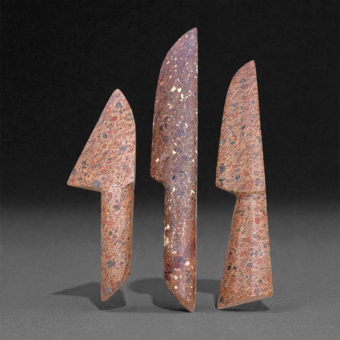 A Group of Three Siberian Fossilized Stone Ritual Knives, 19th Century, 132g, 99AA: Each knife is shaped from fossilized stone with distinct mineral inclusions and tonal variations, ranging from purple-grey to ochre and reddish-brown. The form features a curved, bladed upper edge, a