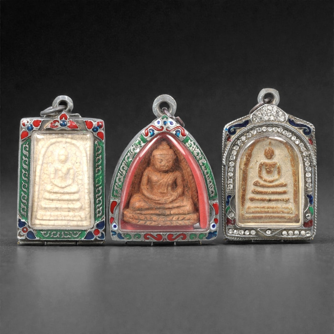 A Group of Thai Clay Buddhist Amulets, 20th Century, 17ZG: Comprising three molded Buddhist amulets formed from pale and reddish-brown clay compositions, each depicting a seated Buddha in meditation posture upon a tiered base. The figures are rendered in low