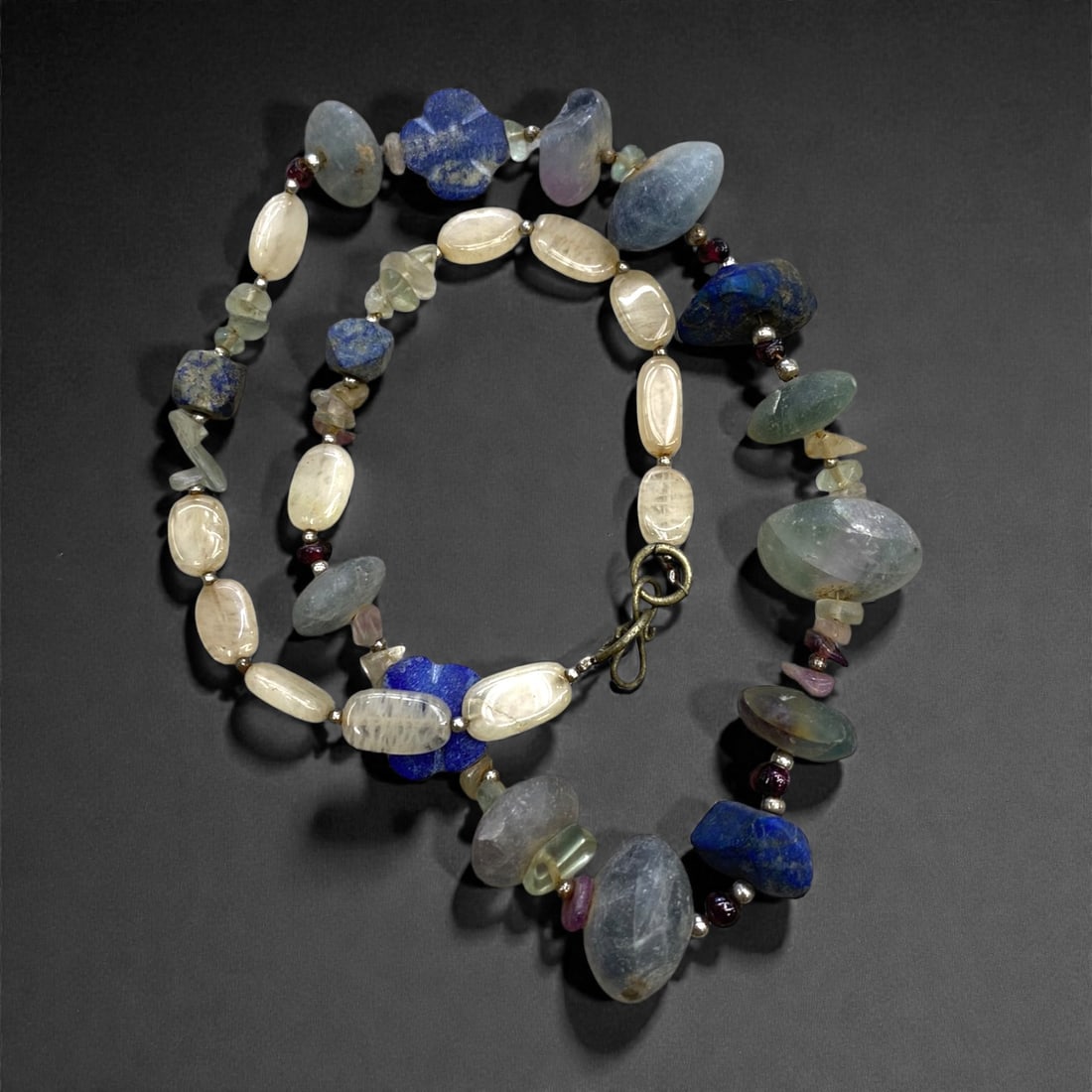 An Ancient Near Eastern Mixed Gemstone Bead Necklace, Bronze Age, 71g, 25AA: Composed of irregularly shaped beads of lapis lazuli, aquamarine, and moonstone, interspersed with small garnet spacers, strung on a modern cord with a period-style metal clasp. The use of lapis lazul