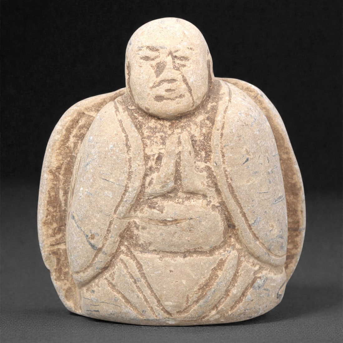 A Japanese Stone Monk Figure, Edo Period, 16ZH: Carved from light beige stone with natural grey and brown patination, the figure depicts a seated monk with hands clasped in a devotional gesture. The head is rounded, with incised lines defining the