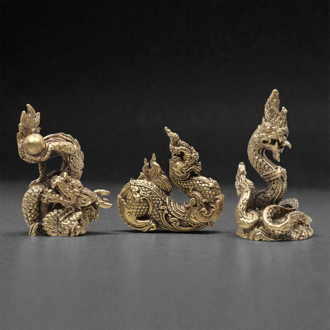 A Set of Three Thai Brass Naga Figures, 20th Century, 67ZG: Cast from brass with a warm golden patina, the set comprises three distinct naga figures. The left figure is coiled, with a spherical element held in its raised coils, detailed scale patterns, and a p