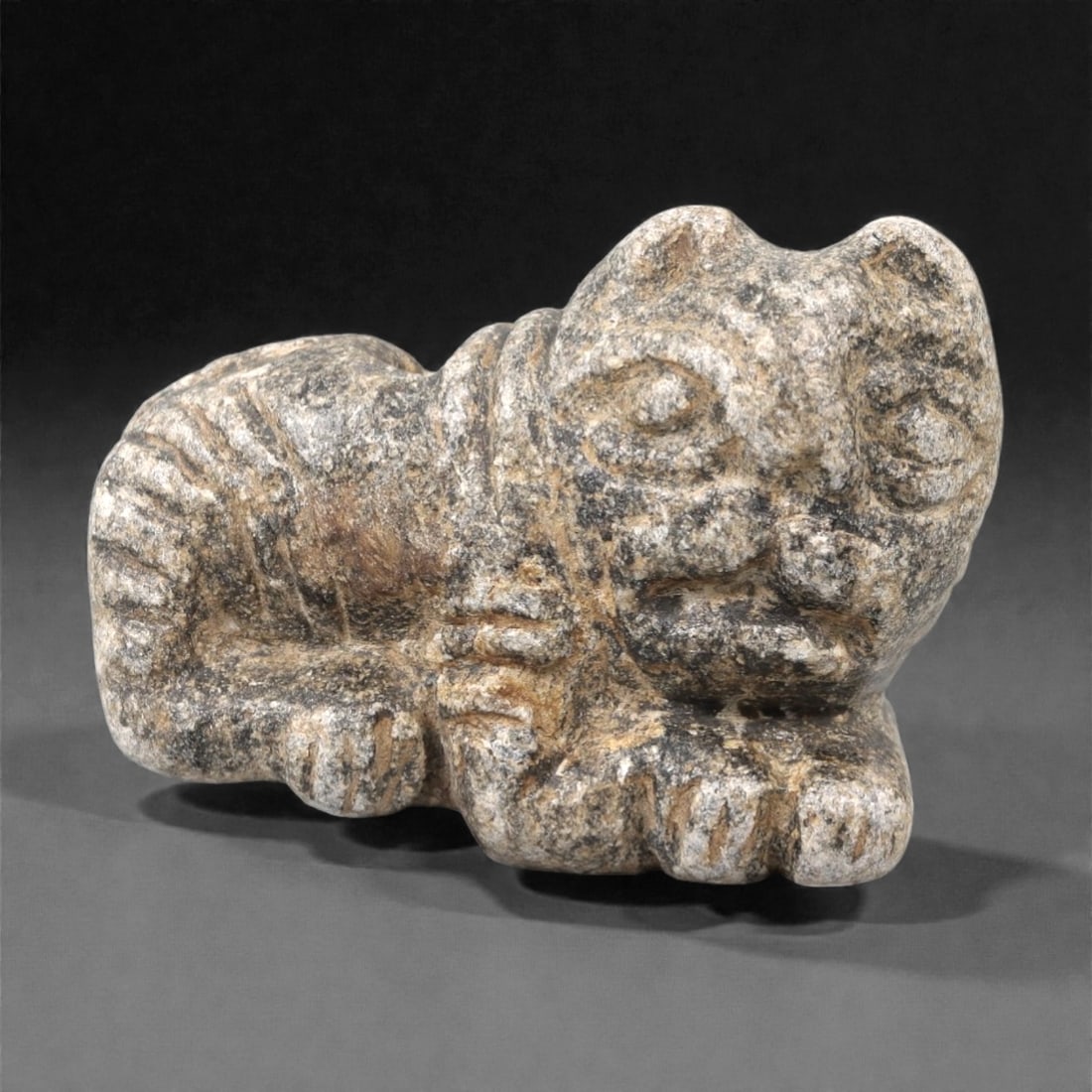 An Ancient Near Eastern Schist Zoomorphic Effigy, 3rd-1st Millennium BCE, 63FL: Carved from grey and white mottled schist with natural mineral inclusions and surface encrustation, the effigy depicts a stylized quadrupedal animal, likely a feline, in a recumbent pose. The head is