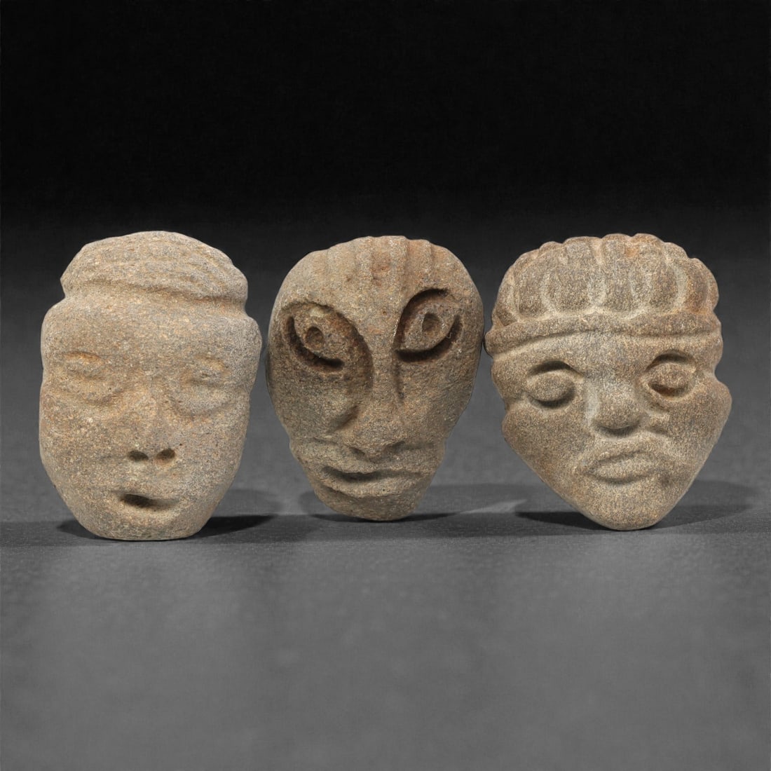 A Group of Three Medieval Stone Maskette Amulets, 10th-14th Century CE, 9ZH: Comprising three distinct maskette amulets, each carved from greyish-brown stone with a granular, weathered surface. The first, of light grey stone, is rounded with closed eyes, a prominent nose, an o
