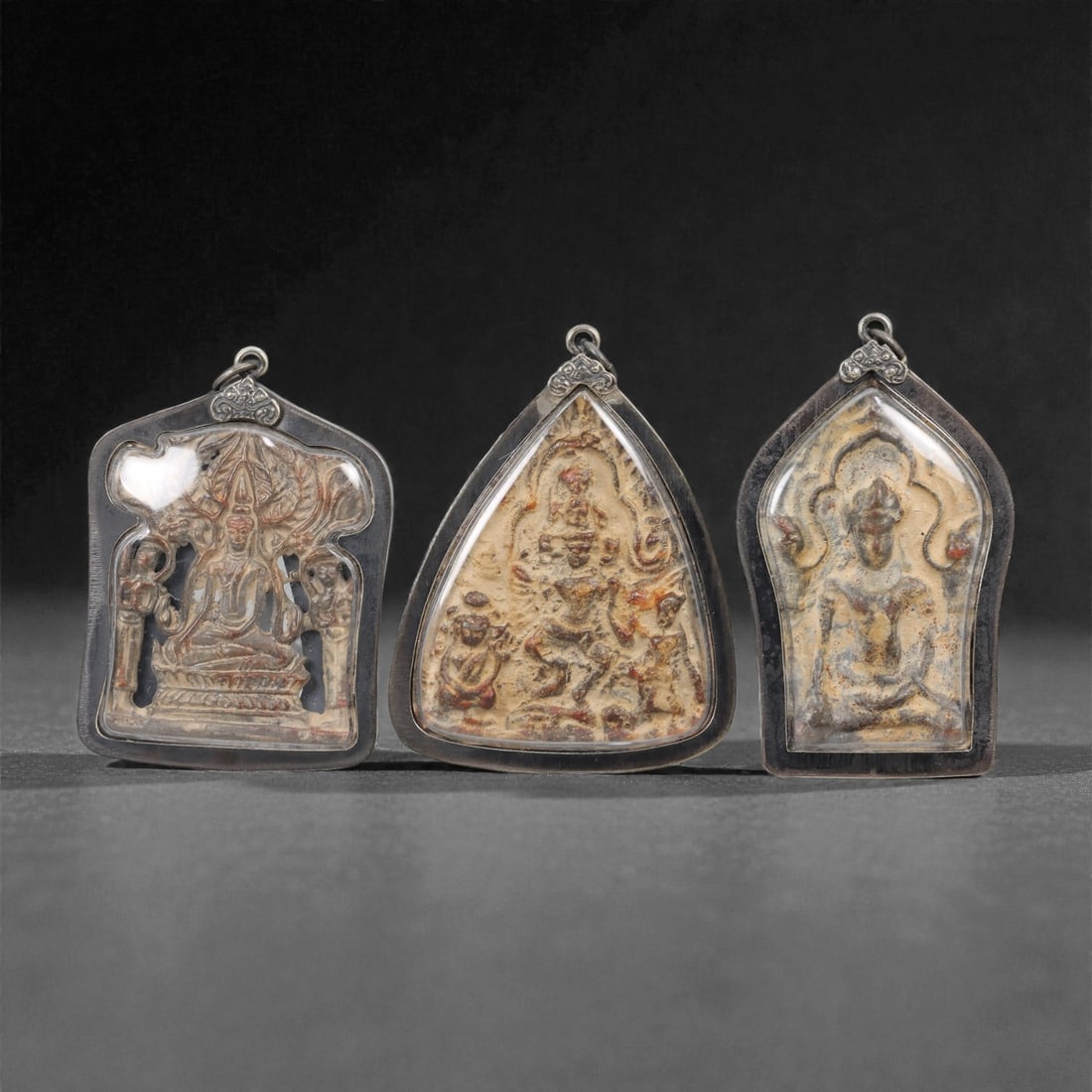 A Group of Three Thai Terracotta Buddhist Amulets, 20th Century, 26FL: Each amulet is formed from terracotta, enclosed in a dark metal case with a suspension loop for wear. The left amulet depicts a seated deity flanked by attendant figures; the central amulet depicts a