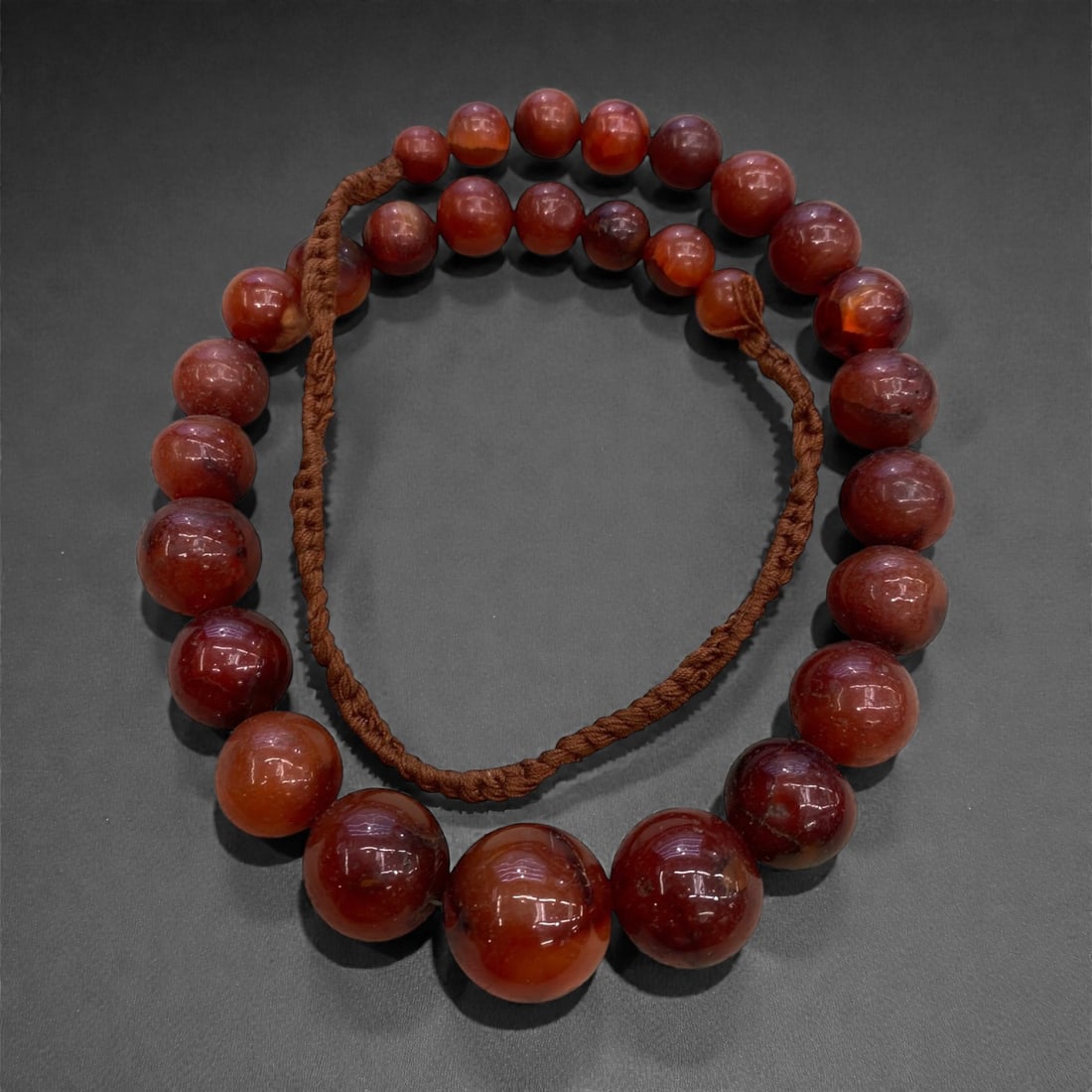 A Near Eastern Style Carnelian Bead Necklace Strand, 19th-Early 20th Century, 155g, 1AA: Composed of graduated spherical carnelian beads ranging in hue from deep reddish-orange to translucent amber, strung on a braided fiber cord. The beads exhibit a smooth, polished surface with natural