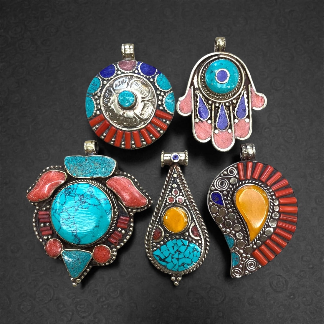 A Group of Five Vintage Silver and Hardstone Inlaid Pendants, 220g, 93AA: Comprising five pendants, each constructed from silver and inlaid with a variety of hardstones. The pendants exhibit distinct forms: a circular example with a central silver floral motif, surrounded b