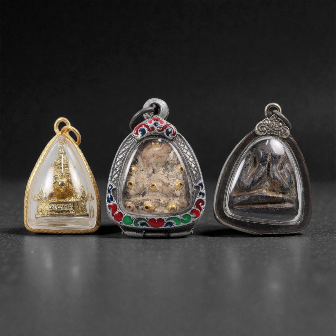 A Group of Three Thai Buddhist Amulets, 20th Century, 24FL: The first amulet is a gilt metal Buddha image, encased in a transparent protective cover with a gold-colored metal frame. The second amulet is composed of a composite material with embedded gold-color