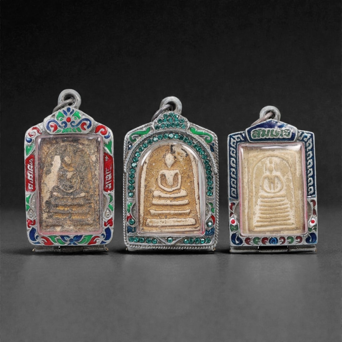 A Group of Three Thai Terracotta Buddha Amulets, 20th Century, 28FL: Each amulet is formed from terracotta, enclosed in a metal case with enamel and glass accents, and fitted with a suspension loop for wear. The amulets depict a seated Buddha in bhumisparsha mudra, wit