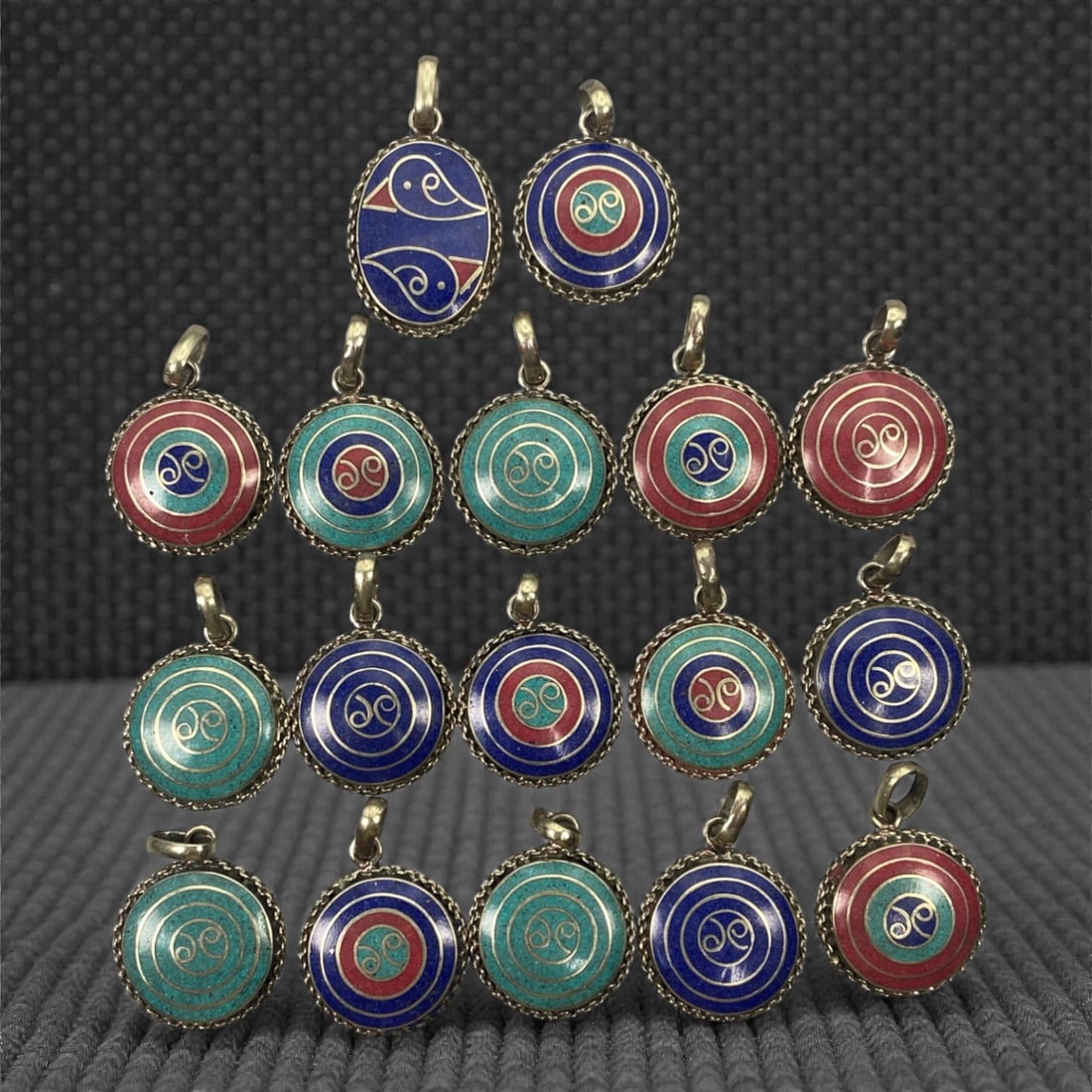A Group of Himalayan Silver and Enamel Pendants, 20th Century, 99FK: The group comprises seventeen small silver-mounted pendants, predominantly circular with one oval example, each fitted with an integral suspension loop. The pendants are decorated with polychrome enam