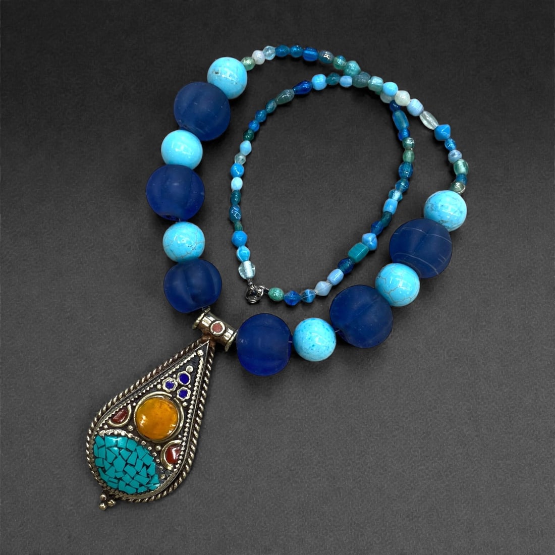 A Central Asian Silver and Mixed Stone Pendant with Glass Bead Necklace, 20thCentury, 123g, 50AA: The necklace is composed of graduated dark blue matte glass beads and polished turquoise composite beads, strung with smaller faceted blue and green glass spacer beads. The pendant is crafted from sil