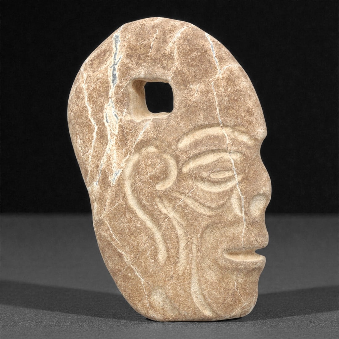 A Pre-Columbian Stone Head Pendant, 10th-15th Century, 11ZH: Carved from light brown stone with natural white mineral veining and visible fissures, the object is shaped as a head in profile with a square suspension hole at the top. The face is rendered in shall