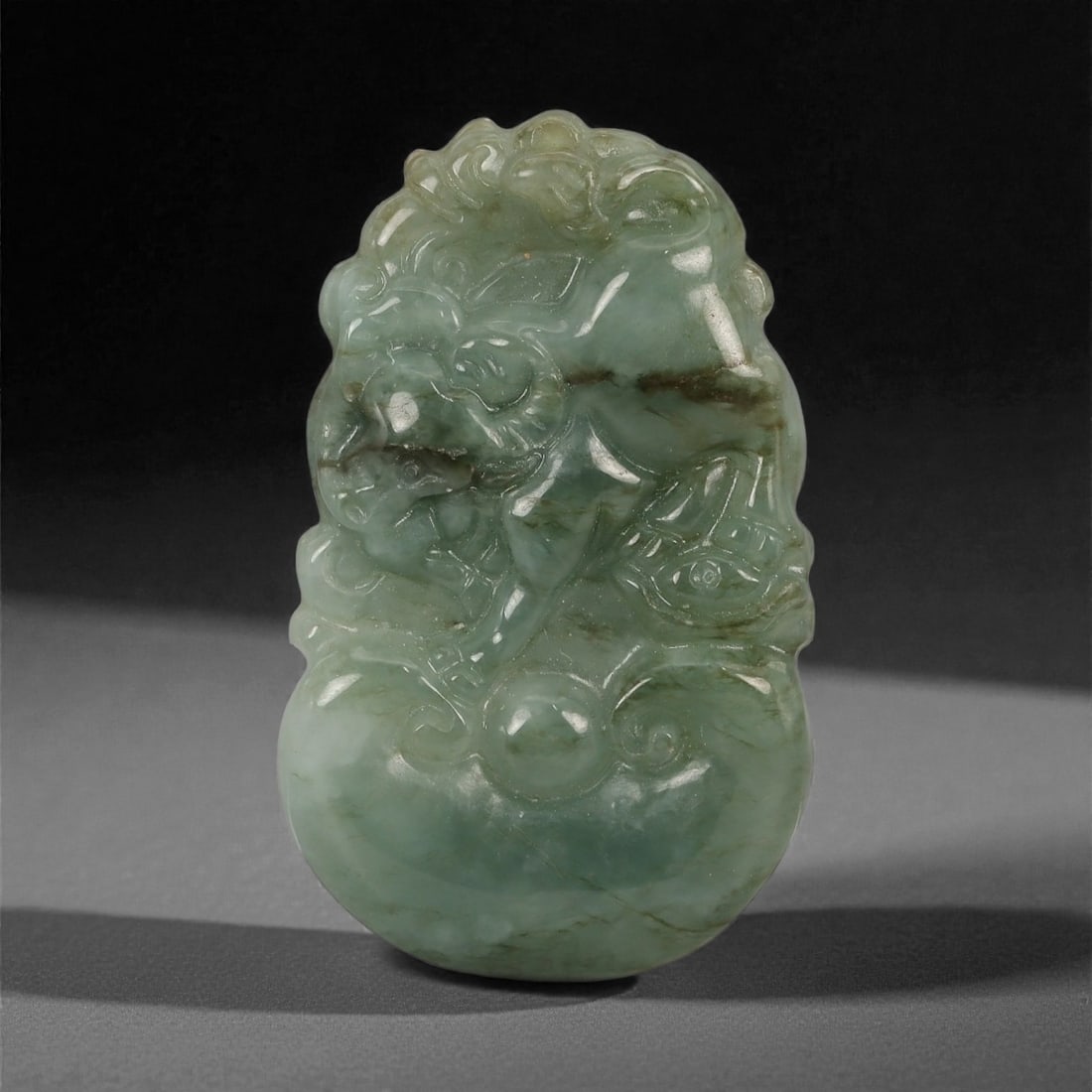 A Chinese Jadeite Jade Bovid Amulet Pendant, 20th Century, 40YB: Carved from mottled green jadeite with areas of darker inclusions and semi-translucent passages, the pendant depicts a bovid shown in a compact standing pose amid stylized cloud scrolls, with the head
