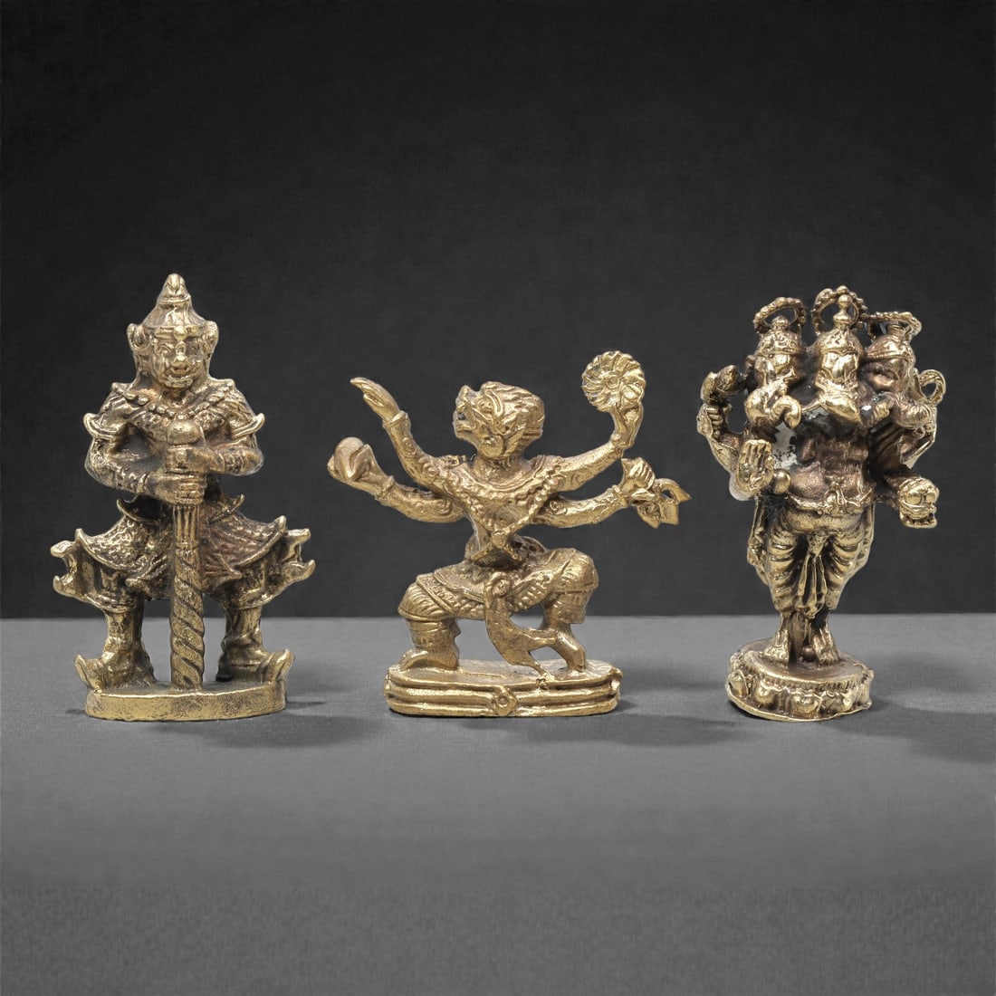 A Set of Three Thai Brass Mythological Figures, 20th Century, 62ZG: Cast from brass with a warm golden patina, the set comprises three distinct mythological figures. The left figure is a guardian-like deity, standing on a rectangular base, clad in ornate armor and hol