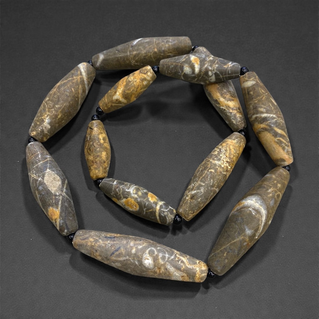 An Ancient Near Eastern Jasper Bead Necklace Strand, 117g, 29AA: Composed of elongated, hand-carved jasper beads in varying earthy tones of brown, grey, and ochre, interspersed with modern black glass spacers, strung on a contemporary cord. These beads exemplify th