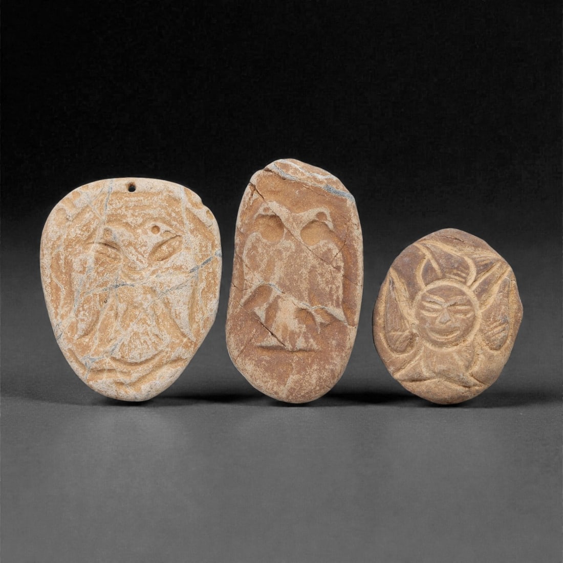 A Group of Three Christian Period Stone Carving Reliefs, 4th-7th Century CE, 52FL: Each relief is carved from light brown stone with natural surface weathering and visible fissures. The first is an ovoid relief depicting a stylized face with incised eyes and a suspension hole at the