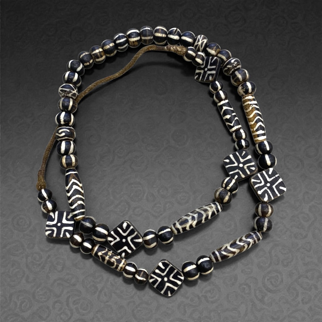 A Southeast Asian Pumtek Wood Bead Necklace Strand, 19th Century, 138g, 12AA: Composed of cylindrical, spherical, and square-shaped wood beads decorated with intricate geometric patterns in black and white, strung on a modern leather cord. The beads are crafted from fossilized