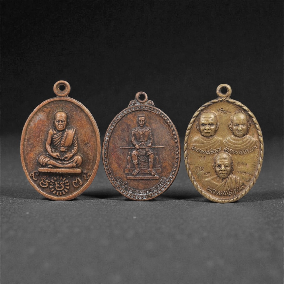 A Group of Three Thai Copper and Brass Amulets, 20th Century, 39FL: Each amulet is cast from copper or brass with a warm patina, shaped as an oval medallion with a suspension loop at the top. The first depicts a seated Buddhist monk in meditative pose, with Khmer scri