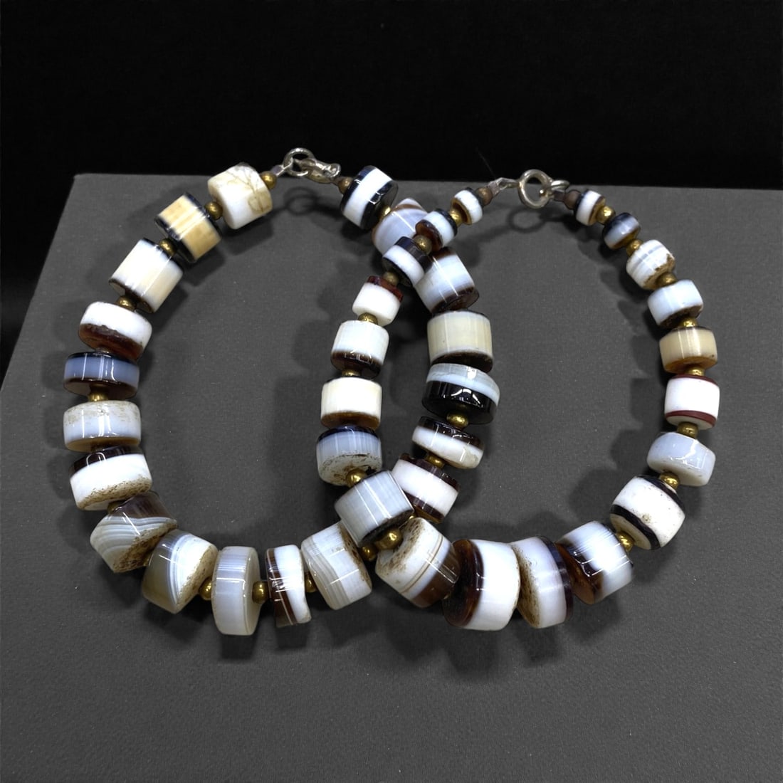 A Group of Two Banded Agate Beaded Necklaces, 45g, 91AA: Comprising two necklaces composed of polished, barrel-shaped banded agate beads, alternating with small gold-toned spacer beads. The beads exhibit concentric banding in contrasting hues of white, brow