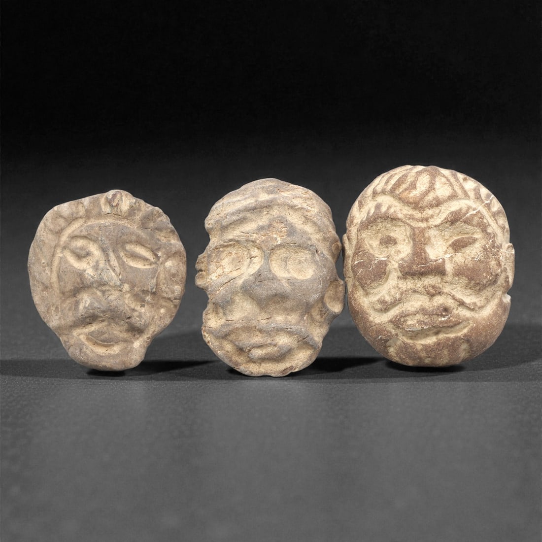A Group of Three Ancient Near Eastern Stone Maskettes, 2nd-1st Millennium BCE, 1ZH: Comprising three distinct stone maskettes, each carved from brownish stone with visible fractures and surface encrustation. The first, of light brown stone, is rounded with closed eyes, a broad nose,