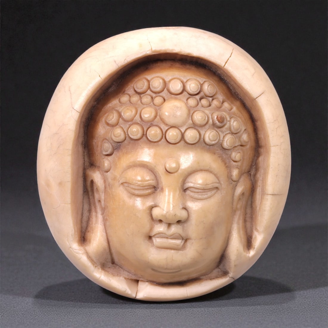 A Chinese Shell Carving of a Buddha Head, 20th Century, 22FC: Carved from a pale, shell-like material with a creamy beige tone and semi-translucent quality, the object represents the head of a Buddha shown frontally within a circular surround. The face is modele