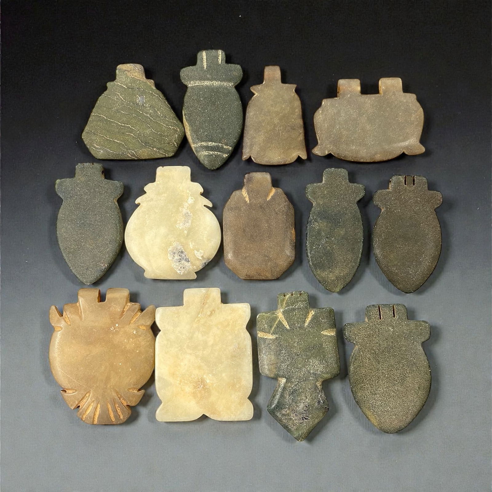 A Group of Eastern Iranian Serpentine Amulets, Islamic Period, 9th-10th Century CE, 56V: Comprising thirteen flat amulets carved from dark green, grey-green, brown, and pale stone, including serpentine and related materials, each formed with a projecting suspension tab and cut in shield-s