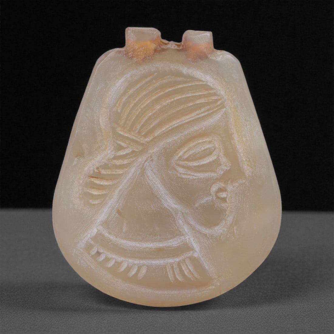 A Mesopotamian Chalcedony Anthropomorphic Amulet, Early 1st Millennium BCE, 73N: Carved from translucent pale honey-colored chalcedony with soft internal clouding and minor surface abrasions, the amulet is formed as a rounded plaque with a flattened reverse and a gently convex fro