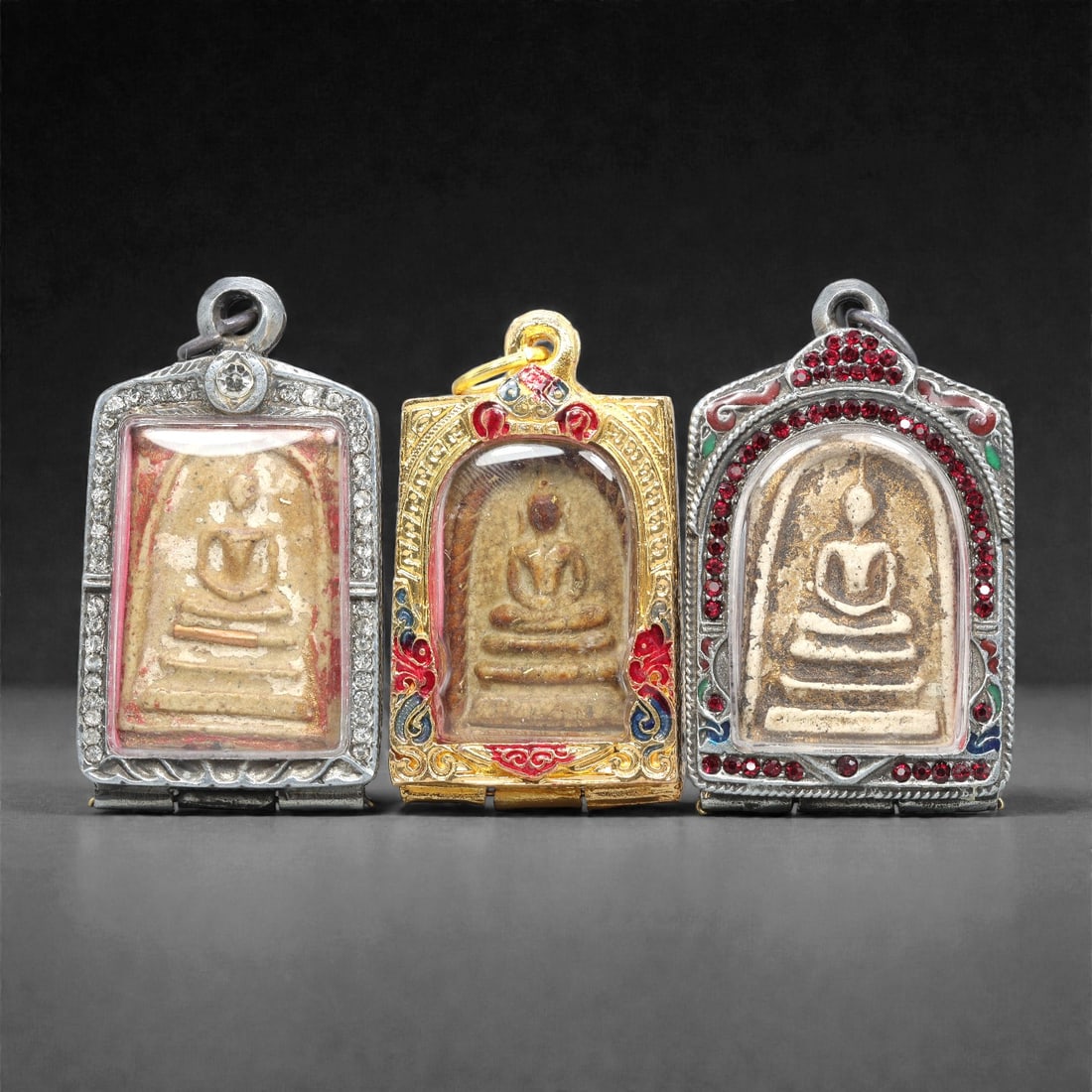A Group of Three Thai Terracotta Buddha Amulets, 20th Century, 84XJ: Each amulet is formed from terracotta, enclosed in a metal case with enamel and glass accents, and fitted with a suspension loop for wear. The amulets depict a seated Buddha in bhumisparsha mudra, wit