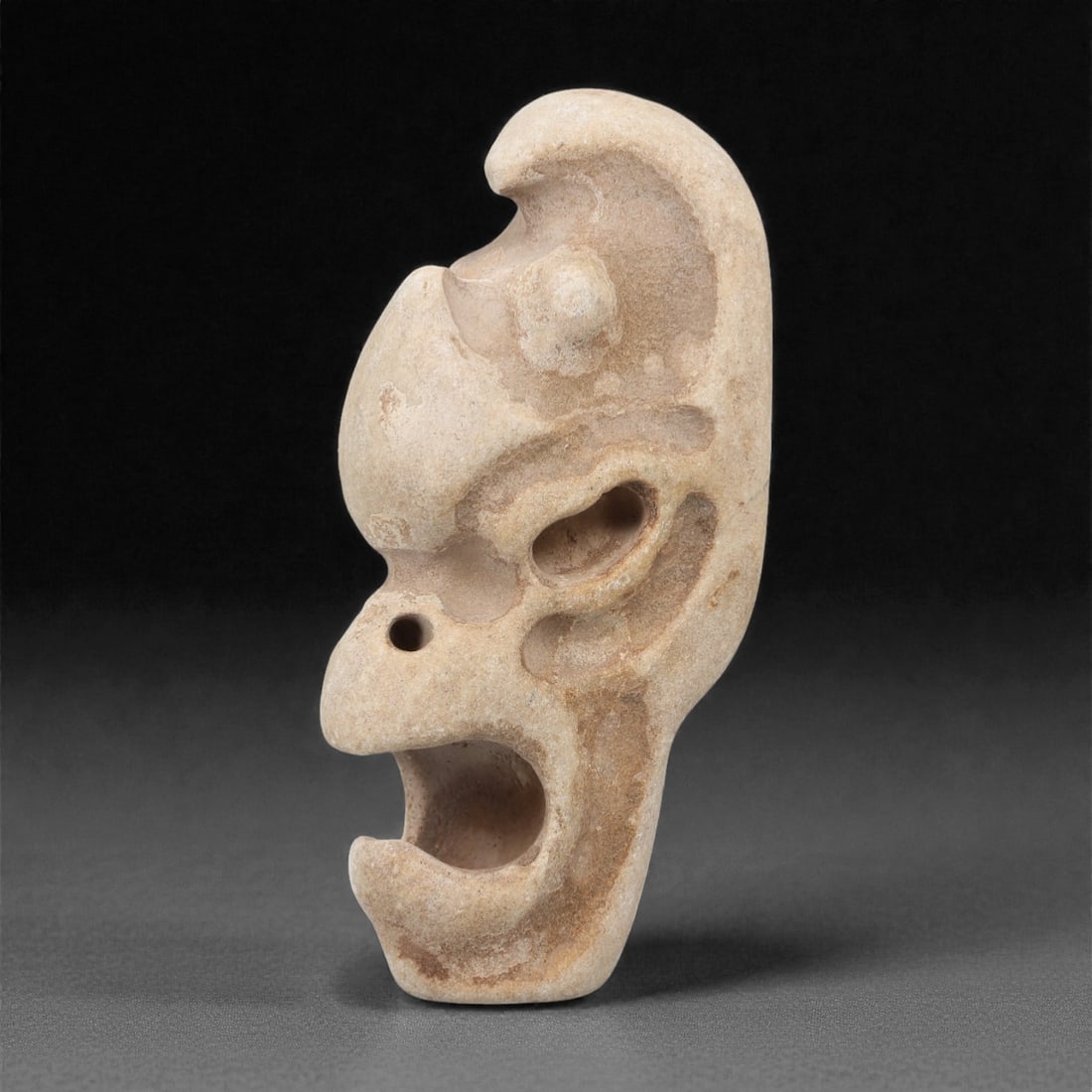 A Pre-Columbian Stone Beast Head, 10th-15th Century, 33FM: Carved from light beige stone with natural patination, the object is shaped as a beast head with a fierce expression, open mouth, and stylized facial features. Incised lines define the eye socket, bro
