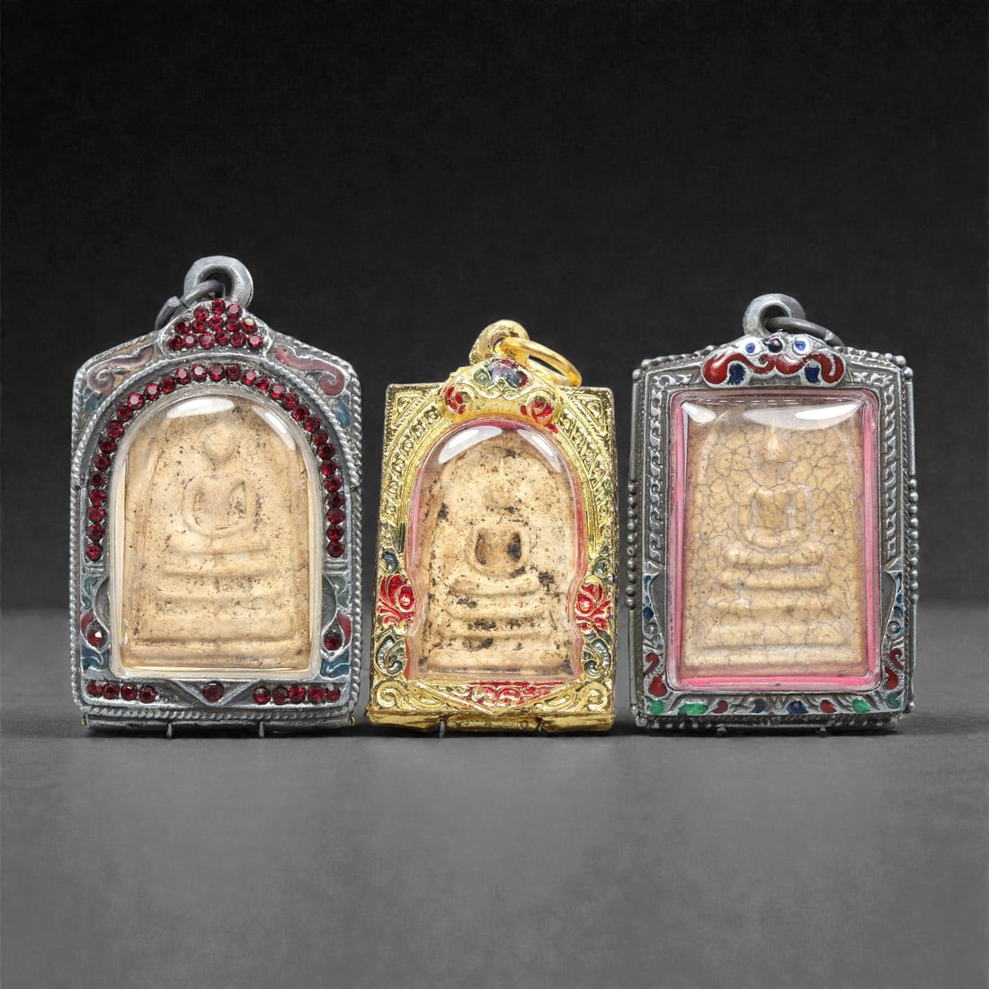 A Group of Three Thai Terracotta Buddha Amulets, 20th Century, 89XJ: Each amulet is formed from terracotta, enclosed in a metal case with enamel and glass accents, and fitted with a suspension loop for wear. The amulets depict a seated Buddha in bhumisparsha mudra, wit