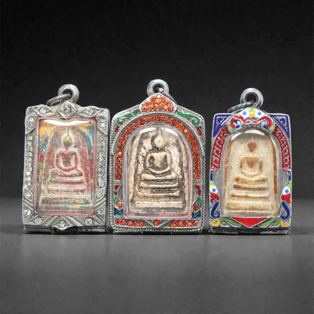 A Group of Three Thai Terracotta Buddha Amulets, 20th Century, 90XJ: Each amulet is formed from terracotta, enclosed in a metal case with enamel and glass accents, and fitted with a suspension loop for wear. The amulets depict a seated Buddha in bhumisparsha mudra, wit