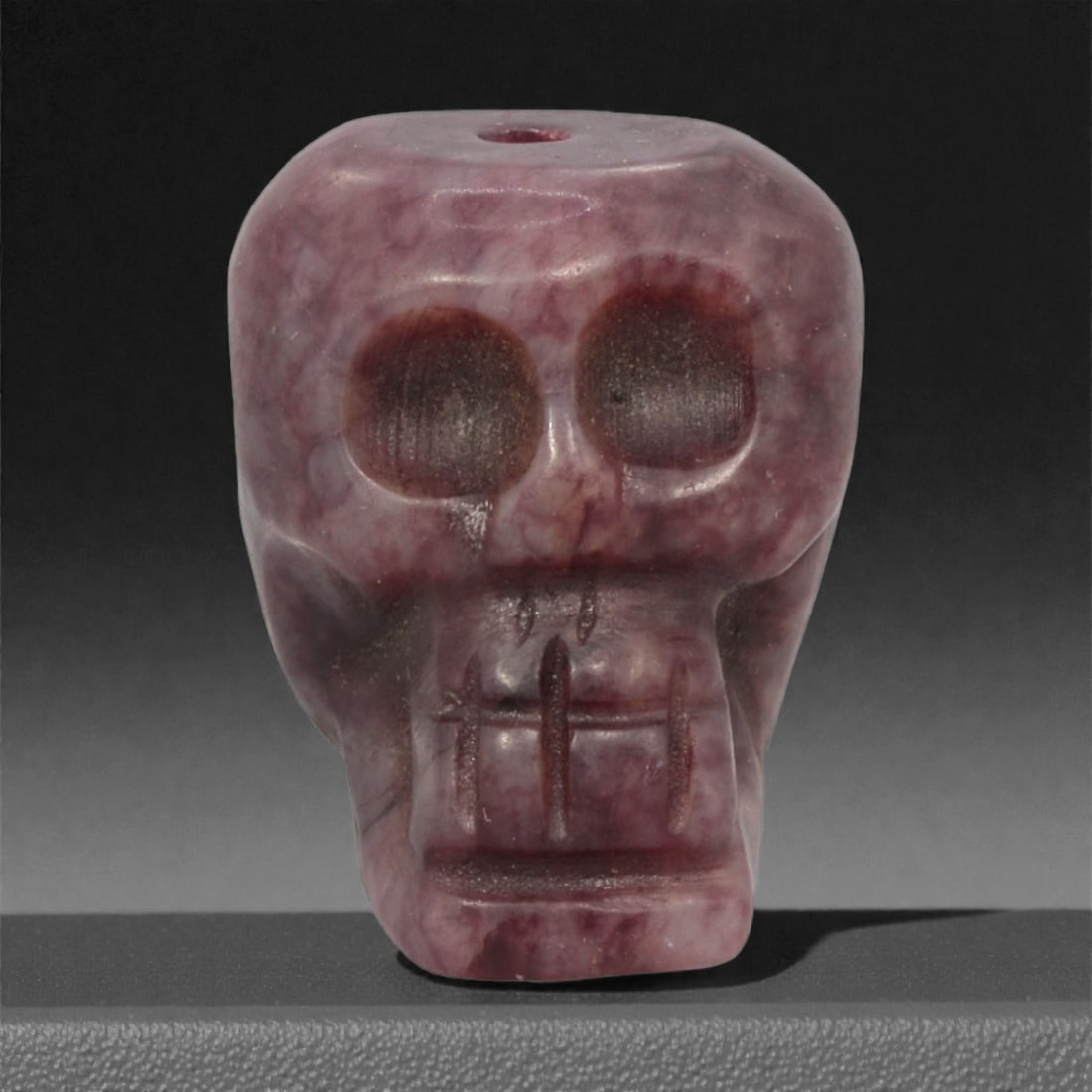 A Tibetan Stone Skeleton Bead, Mid-20th Century, 58FJ: Carved from translucent purplish stone with darker maroon inclusions and subtle internal veining, the bead is formed as a stylized human skull rendered in frontal view, with large circular recessed ey