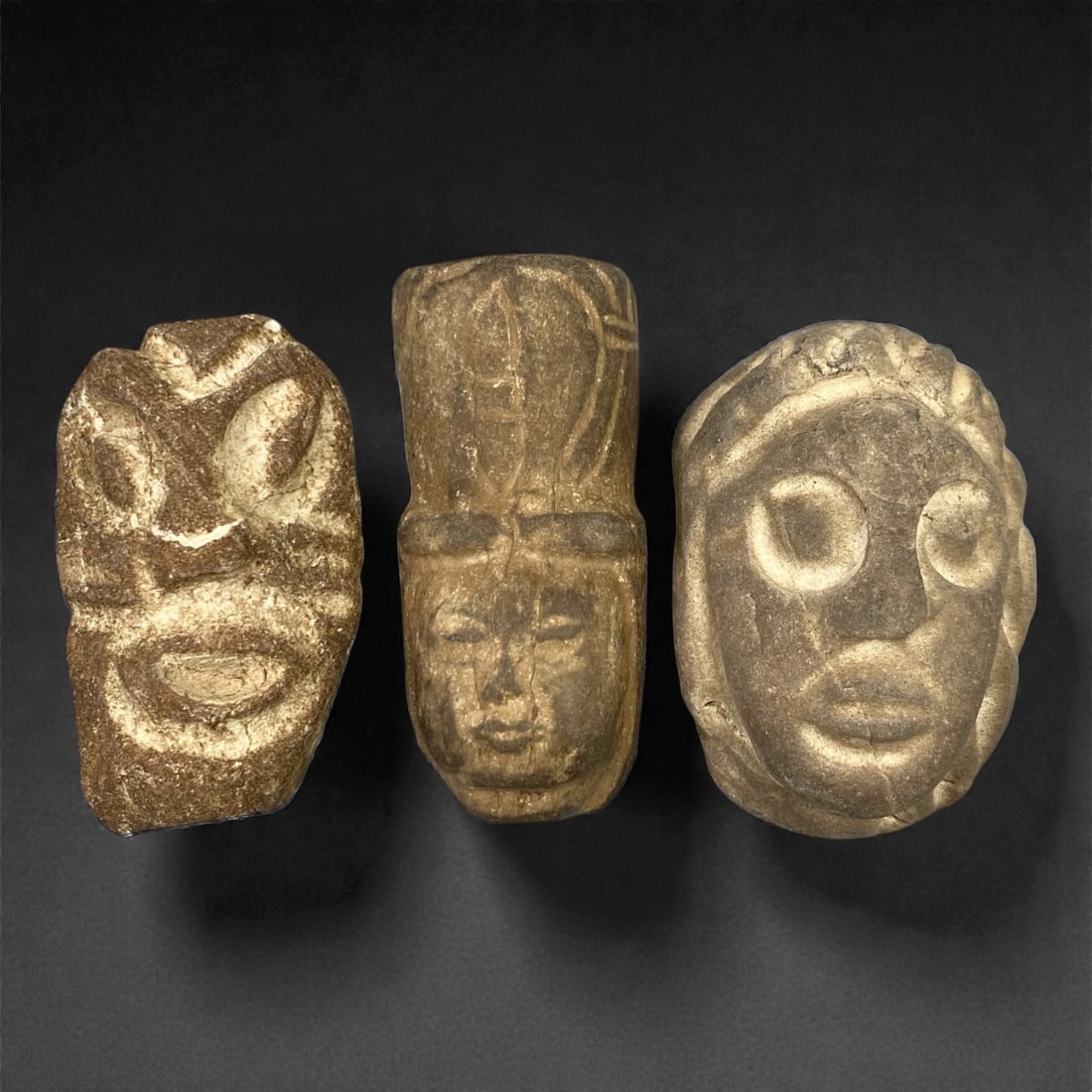A Group of Three Ancient Stone Maskettes, 1st Millennium BCE, 77FM: Comprising three stone maskettes, each formed from light brown to grey stone with visible surface fissures and tonal variation. The leftmost maskette features a stylized, expressive face with recessed