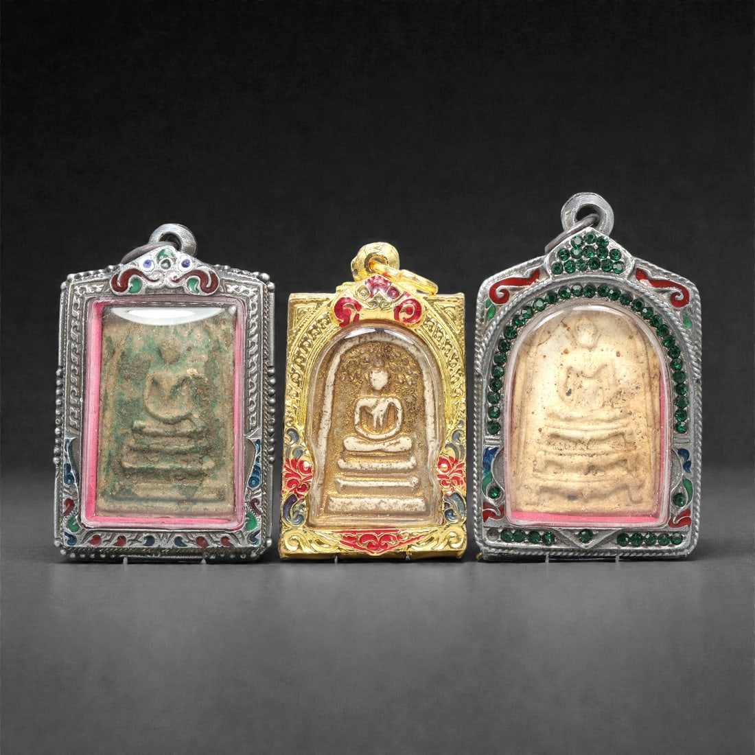 A Group of Three Thai Terracotta Buddha Amulets, 20th Century, 82XJ: Each amulet is formed from terracotta, enclosed in a metal case with enamel and glass accents, and fitted with a suspension loop for wear. The amulets depict a seated Buddha in bhumisparsha mudra, wit