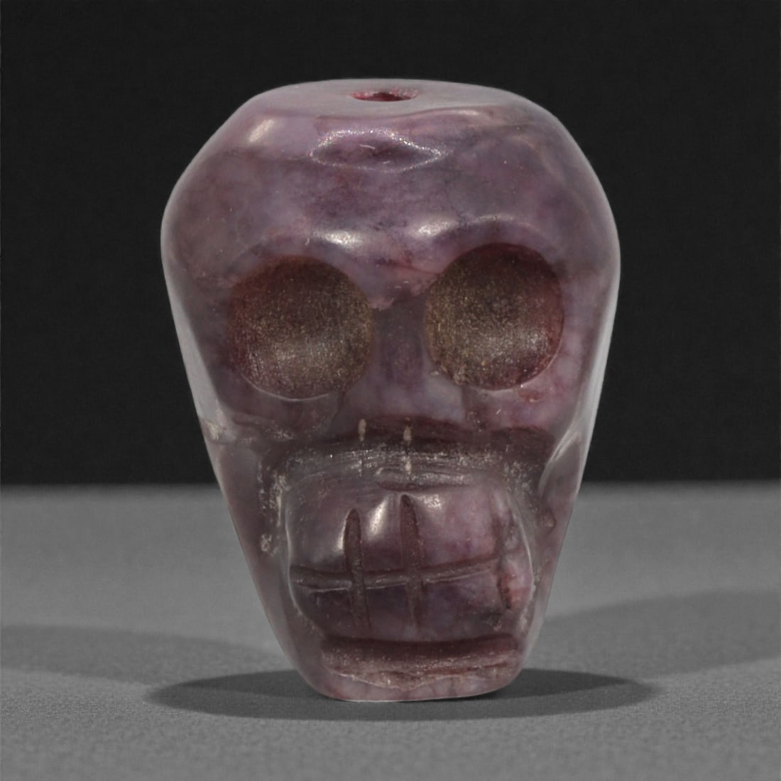 A Tibetan Stone Skeleton Bead, Mid-20th Century, 60FJ: Carved from translucent purplish stone with darker brown inclusions and natural internal veining, the bead is formed as a stylized human skull rendered in frontal view, with large circular recessed ey