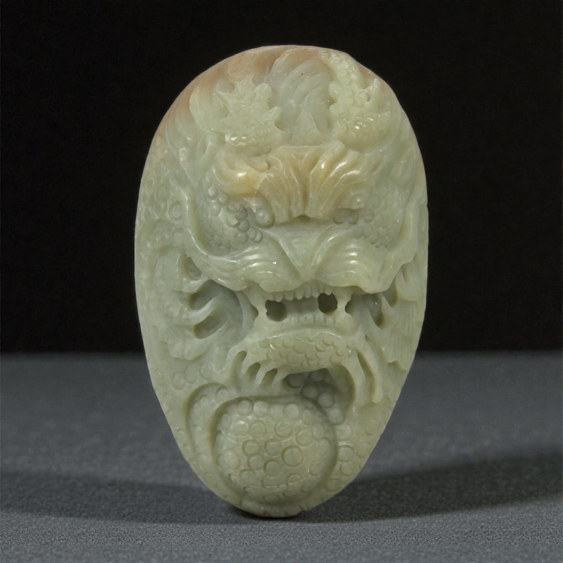 A Chinese Jade Dragon Mask Pendant, 20th Century, 98YA: Carved from pale celadon jade with areas of creamy translucency and subtle natural inclusions, the pendant is worked in high relief as a frontal dragon mask. The face is powerfully modeled with promin