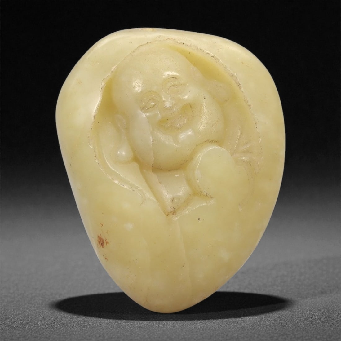 A Chinese Hetian Jade Pebble Carving of Budai, 20th Century, 92FH: Carved from a naturally rounded Hetian nephrite pebble of pale yellowish celadon tone, the carving depicts Budai shown in relief emerging from the natural contours of the stone. The figure is rendered