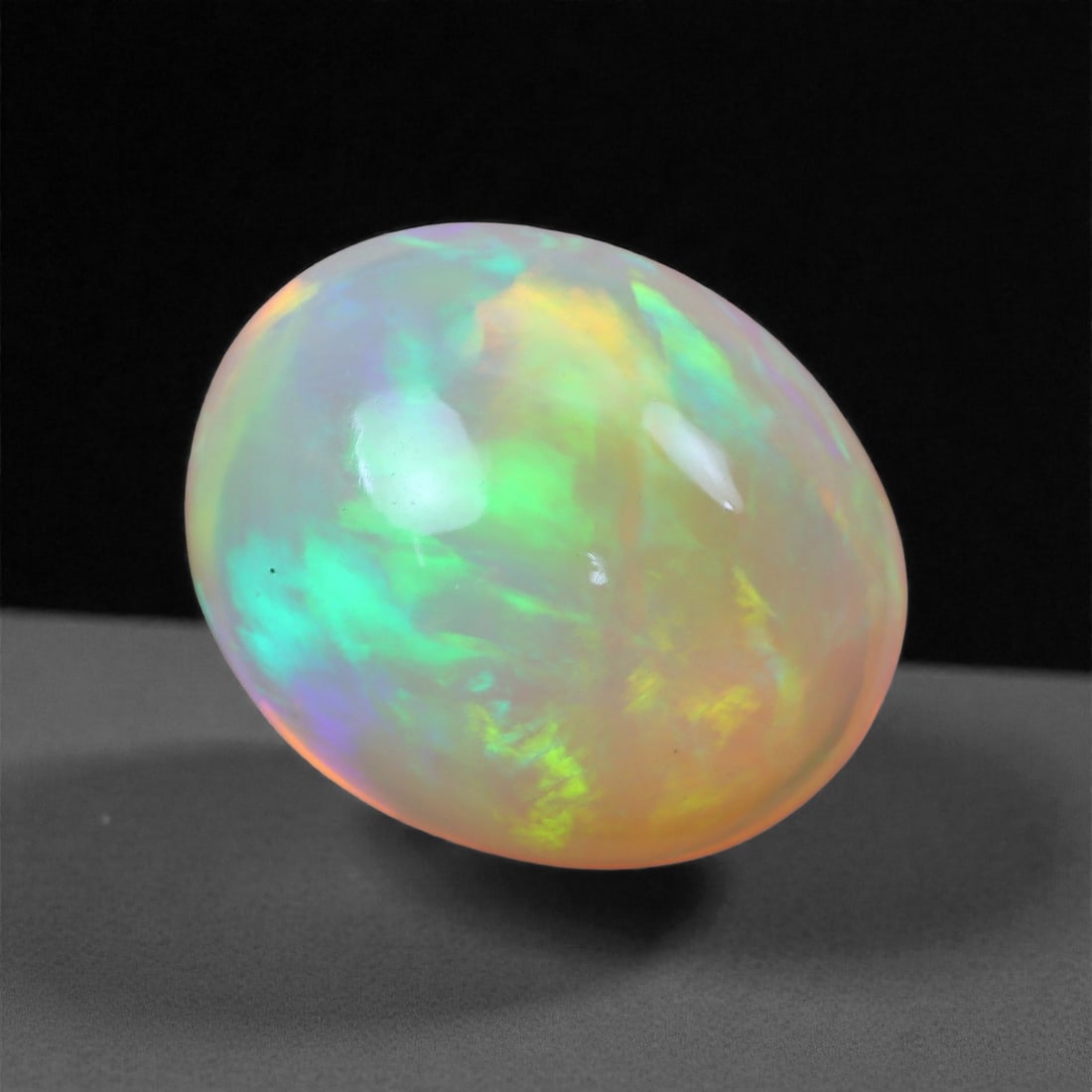 3.33 Carats Oval Cabochon Opal - Natural Ethiopian Stone 11.1x9.6x6.6mm, 44XH: This vibrant Ethiopian opal bursts with brilliant neon greens, electric yellows, and hints of violet flashing beneath its polished dome. The gemstone's compact oval shape and translucent body make the