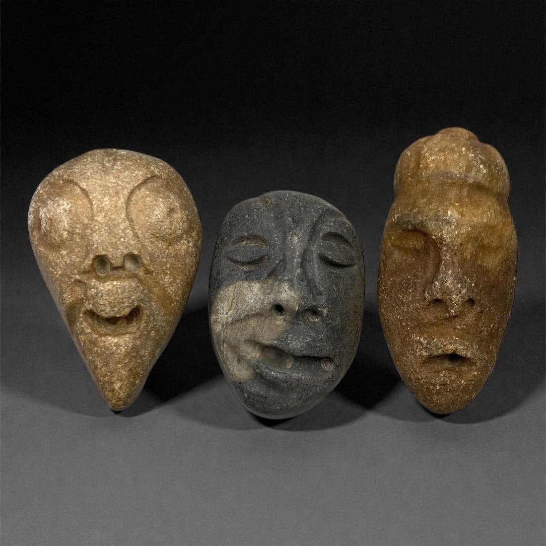 A Group of Three Medieval Stone Maskette Amulets, 10th-14th Century, 11FM: Carved from stone with varied natural patination, the set comprises three miniature maskette amulets: one in light tan stone with prominent circular eyes and a pointed base, one in dark grey stone wit